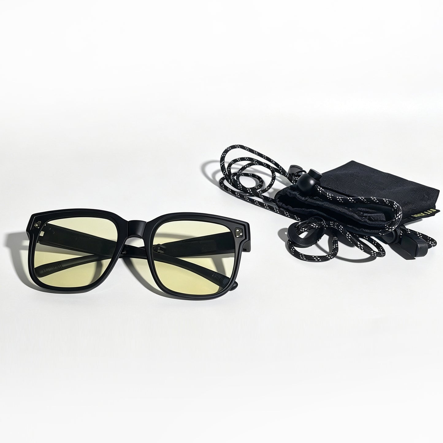 Matte black fit-over night driving glasses with yellow lenses beside a storage pouch and adjustable neck strap.