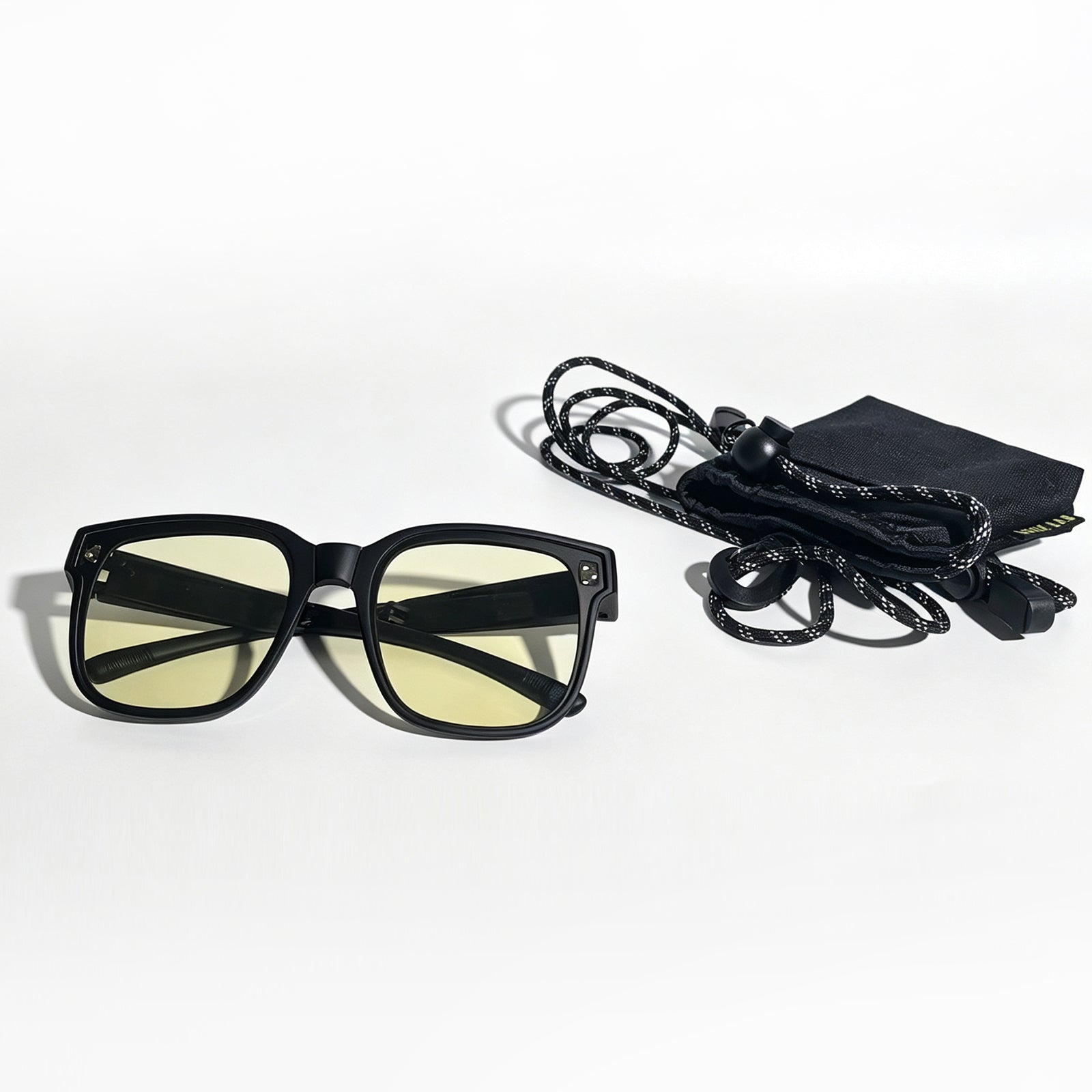 Matte black fit-over night driving glasses with yellow lenses beside a storage pouch and adjustable neck strap.